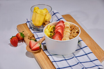 A tropical fruit bowl filled with fresh mango, strawberry, and coconut flakes, served in a white cup on a wooden board. Surrounded by whole mango, watermelon slice, strawberries, and granola.