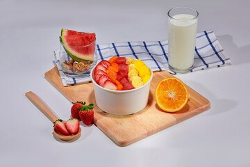 A tropical fruit bowl filled with fresh mango, strawberry, and coconut flakes, served in a white cup on a wooden board. Surrounded by whole watermelon slice, strawberries, orange.