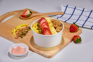 A tropical fruit bowl filled with fresh mango, strawberry, and coconut flakes, served in a white cup on a wooden board. Surrounded by whole mango, watermelon slice, strawberries, and granola.