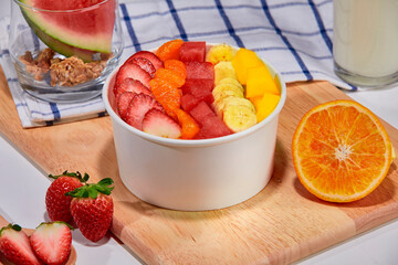 A tropical fruit bowl filled with fresh mango, strawberry, and coconut flakes, served in a white cup on a wooden board. Surrounded by whole watermelon slice, strawberries, orange.