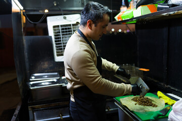 Cook adding savory meat to a tortilla in a bustling food truck at night, skillfully preparing...
