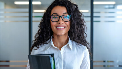 Confident and businesslike young beautiful girl with bouffant and curly hair, smiling in the office, wearing glasses and a white shirt