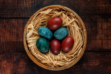 A small collection of red and blue dyed eggs placed in a basket lined with straw, resting on a dark wooden background. The warm lighting and natural textures create a cozy and nostalgic atmosphere.