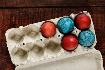 A simple yet artistic arrangement of red and blue dyed eggs in a recycled carton on a dark wooden table. The contrast of colors and textures highlights the natural imperfections and beauty.