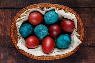 A basket filled with blue and red dyed eggs, carefully placed on a neutral fabric lining, sitting on a dark wooden surface. The interplay of textures and tones creates a harmonious and warm visual.