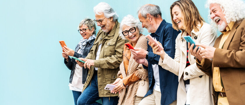 Group of senior people using technology devices together standing on a blue wall - Happy older friends having fun watching funny video on smartphone - Tech and modern elderly concept	