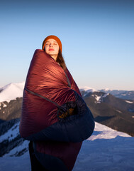 Young woman with closed eyes, standing in front of a winter mountain, wrapped in a warm sleeping...