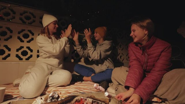 Joyful friends gathering around campfire, playing hand-clapping games, roasting marshmallows during nighttime outdoor bonding experience