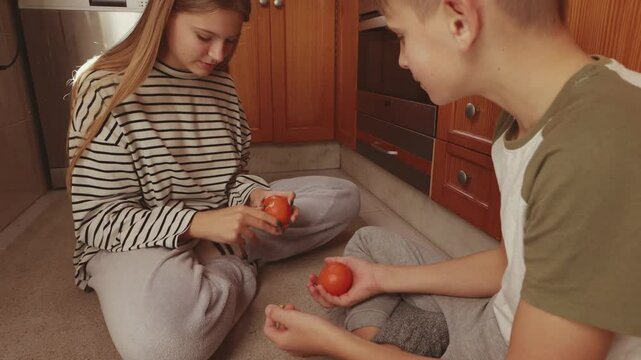 Teenage siblings resting on kitchen floor, peeling mandarin, sharing intimate moment of familial closeness and casual connection