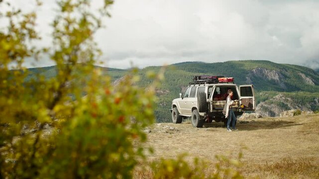 Woman wearing a wool cardigan pouring pancake batter into a pan on a camping stove set up on the tailgate of an off-road vehicle, enjoying a peaceful outdoor cooking experience