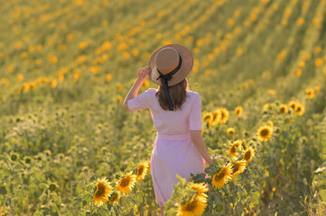 Ein M&auml;dchen in einem Hut und einem pinken Kleid steht mit dem R&uuml;cken in einem Sonnenblumenfeld und schafft eine warme und malerische Szene.
