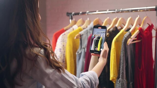 Young influencer creating video content for local store with fashion clothes, woman using smartphone for filming garment assortment and then uploading for channel promotion, social media content