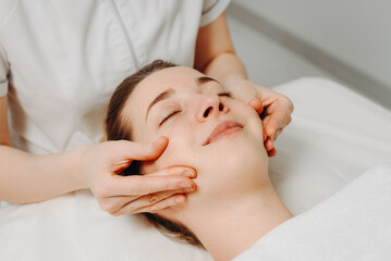 face massage, young attractive caucasian woman, beautiful relaxed lady, white towel, spa therapist doing facial and head massage in beauty salon, massaging cheeks, skincare and bodycare concept