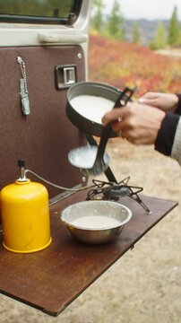 Woman wearing a wool cardigan pouring pancake batter into a pan on a camping stove set up on the tailgate of an off-road vehicle, enjoying a peaceful outdoor cooking experience