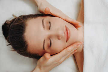 face massage, young attractive caucasian woman, beautiful relaxed lady with closed eyes, view from above, spa therapist doing facial and head massage in beauty salon, skincare and bodycare concept