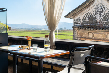 A serene outdoor café with a stunning countryside view, featuring a table set with a glass bottle of lemon water, a drinking glass, and a small flower vase. 