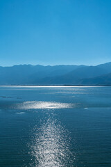 A breathtaking view of a vast lake with shimmering blue waters, surrounded by majestic mountain ranges under a clear sky. The sunlight reflects beautifully on the water’s surface, 
