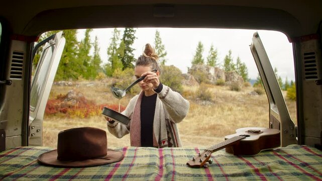 Woman wearing a wool cardigan pouring pancake batter into a pan on a camping stove set up on the tailgate of an off-road vehicle, enjoying a peaceful outdoor cooking experience