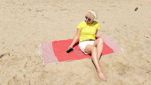 Beautiful cheerful sixty year old blonde woman listening to music in big white headphones sitting on the beach. Woman in yellow T-shirt and white shorts. Pensioner woman in sunglasses