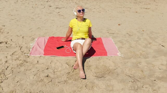 Beautiful cheerful sixty year old blonde woman listening to music in big white headphones sitting on the beach. Woman in yellow T-shirt and white shorts. Pensioner woman in sunglasses