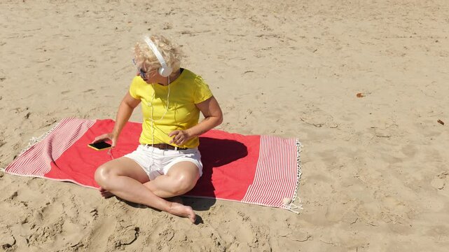 Beautiful cheerful sixty year old blonde woman listening to music in big white headphones sitting on the beach. Woman in yellow T-shirt and white shorts. Pensioner woman in sunglasses