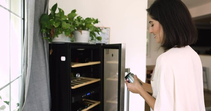 Positive beautiful young black haired woman taking can of cold drink from small minibar fridge, using facilities in rental accommodation, hotel room, modern home appliance technology