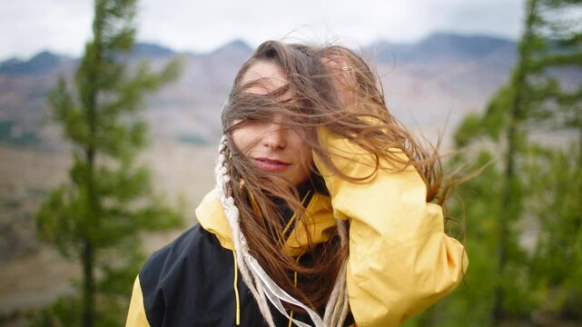 Young woman hiking in a stunning mountain landscape, holding her hair back against the wind. Dressed in a bright yellow jacket, she sports braids surrounded by coniferous trees