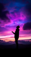 Fototapeta premium Silhouette of person standing with outstretched hand against vibrant purple and pink sunset sky, mountains in background creating spiritual, peaceful atmosphere.