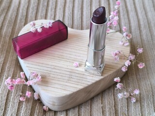 burgundy lipstick in a silver case on a wooden heart and gypsophila flowers beige background