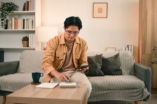A man in casual attire sits on a cozy sofa, using a calculator and a smartphone, representing financial planning, budgeting, and expense tracking, Home Finance Management concept