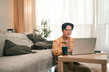 An Asian adult male sits on the floor, working on his laptop in a cozy home, enjoying the flexibility of Remote work made easy for the modern digital nomad