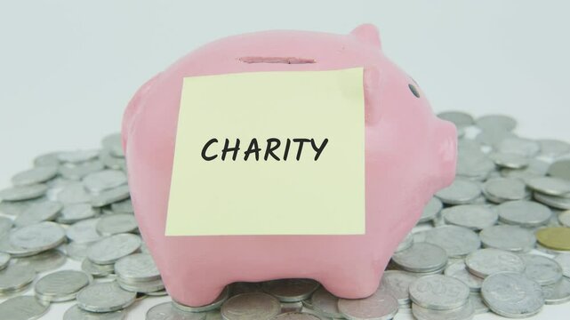 Charity text on a yellow note with a hand inserting coins into a piggy bank, surrounded by a pile of coins on a white background, symbolizing generosity and donations