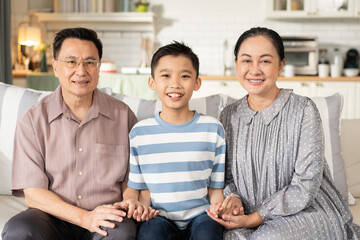 Happy Asian grandson and grandparents relax sitting on couch in the living room looking at camera. Family generation love bonding, people smiling and laughing together at home