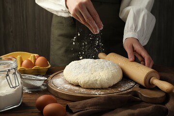 Woman with rolling pin adding flour onto dough at wooden table, closeup