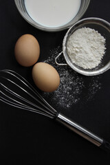 fresh eggs, milk in a plate, flour in a sieve and scattered on the background, a whisk lies on a dark background, different angles
