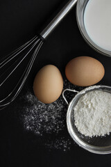 fresh eggs, milk in a plate, flour in a sieve and scattered on the background, a whisk lies on a dark background, different angles
