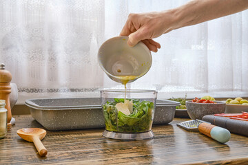 Hand pours lemon juice into a food processor filled with fresh green herbs and garlic
