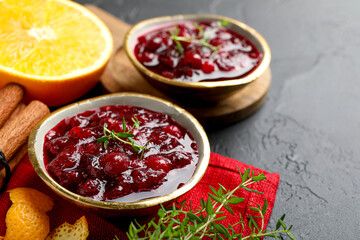 Tasty cranberry sauce in bowls, orange, thyme and cinnamon on black table, closeup