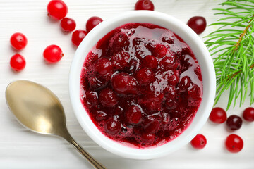 Tasty cranberry sauce in bowl, berries, spoon and fir tree branches on white wooden table, top view