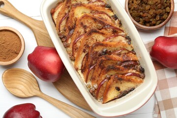 Delicious bread pudding in baking dish, fresh apples, cinnamon and raisins on white table, flat lay