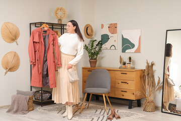 Fototapeta premium Young woman with stylish coat near clothes rack at home