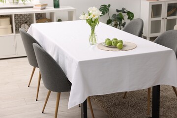 Table with white tablecloth, apples, flowers in vase and chairs in stylish room. Interior design