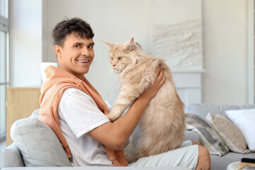Handsome young happy man with cute Maine Coon cat sitting on sofa at home