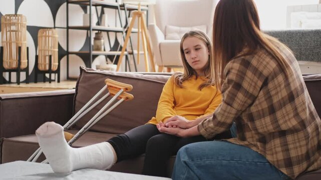 Girl with broken leg in cast sits with mother on couch at home. Sad little kid hugging teddy bear recovering from broken leg. Girl using cast crutches for quick rehabilitation. Mom supporting daughter