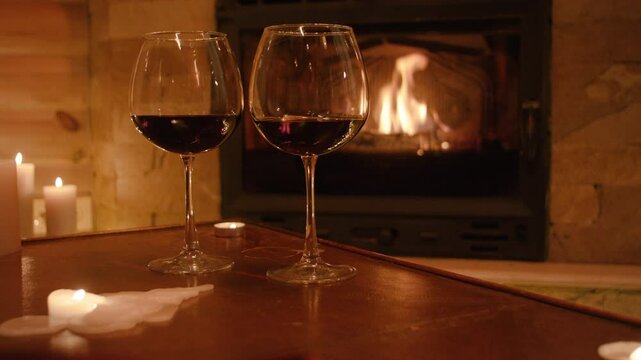 Two wine glasses and fading candles rest on a table, one dripping wax across the surface. A glowing fireplace in the background and scattered candlelight create a warm, intimate ambience.