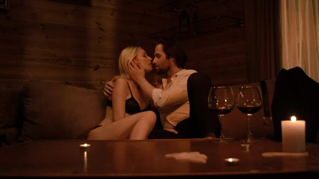 A blonde in lingerie reclines on a beige sofa as a brunette man in a white shirt and black pants gently caresses her neck and face. Three candles and two wine glasses on the table set a sultry mood.