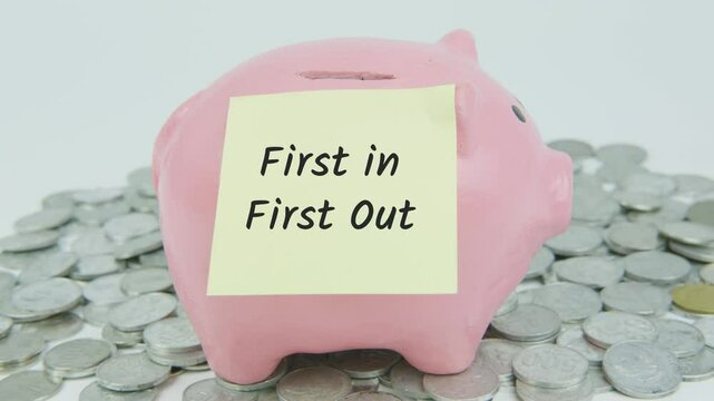 FIFO First In First Out written on a yellow note paper with a hand inserting coins into a pink piggy bank, surrounded by a pile of coins on a white background