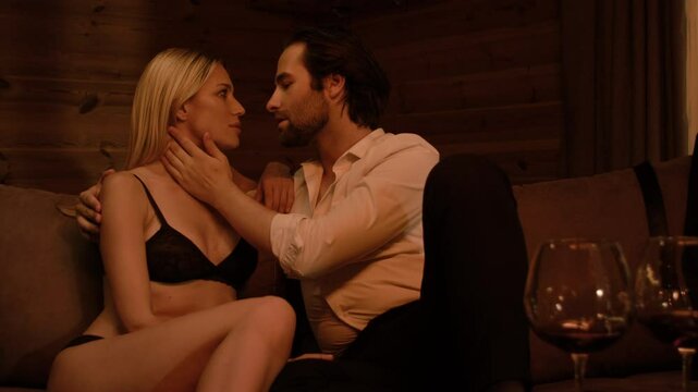 A stubbled brunette man in a white shirt and black pants gently caresses a blonde in black lingerie, their faces pressed close, lips almost touching. Two wine glasses complete the scene.