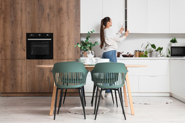 Young woman taking glass from cupboard in kitchen