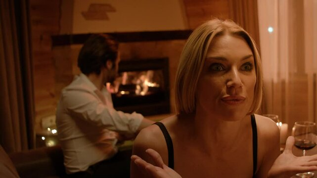 A blonde in black lingerie sits forward, eyes wide and lips parted in passionate speech. Her raised hand emphasizes emotion, while a brunette man by the fireplace turns away under soft candlelight.